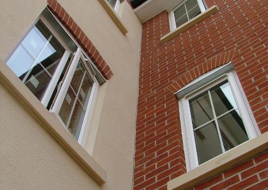 Side profile of a red brick new build home. You can see two stories height of building, and the windows are simple white double glazed windows slightly ajar. The sky above the guttering and roof is bright blue.