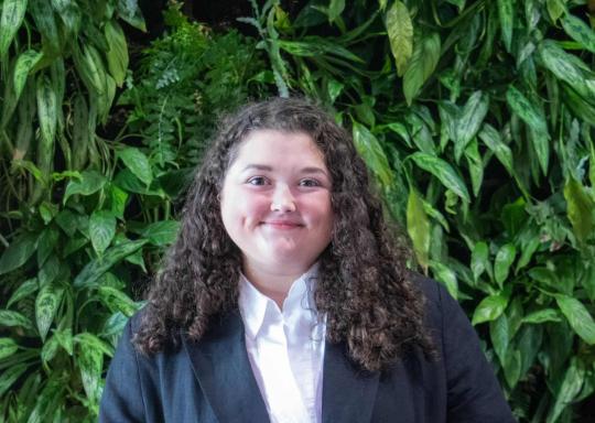 Bethany Bale, Policy and Campaigns Officer, stands smiling in front of a wall of green leaves