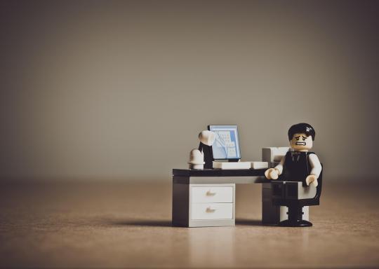 Image shows a white, grey and black desk with a man with a worried look on his face seated on a black chair looking directly at the camera. The desk, man and chair are all made of lego bricks.